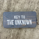 Made in the UK by Giggle Gift Co. Wooden block keyring with white text quote on both sides; 'Key to the Unknown'  grey