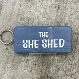 Made in the UK by Giggle Gift Co. Wooden block keyring with white text quote on both sides; 'The She Shed'  grey