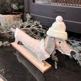 Smiling Dachshund in felt 34cm wearing a white bobble hat and a cream sparkling dog coat with faux fur edging stood on a wooden plinth