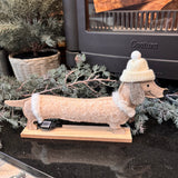 Smiling Dachshund in felt 34cm wearing a white bobble hat and a cream sparkling dog coat with faux fur edging stood on a wooden plinth