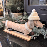 Smiling Dachshund in felt 34cm wearing a white bobble hat and a cream sparkling dog coat with faux fur edging stood on a wooden plinth