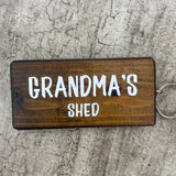 Made in the UK by Giggle Gift Co. Wooden block keyring with white text quote on both sides; 'Grandma's Shed'  brown