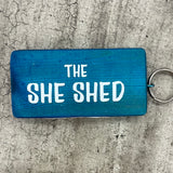 Made in the UK by Giggle Gift Co. Wooden block keyring with white text quote on both sides; 'The She Shed'  blue
