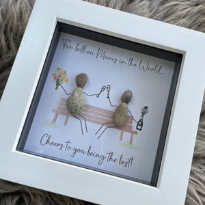 Mini Framed Pebble Art - White block square frame 12.5cmWith a soft background image of a bench and two pebble people sitting on it celebrating with a champagne toast and a bunch of flowers with the quote 'Two Billion Mums in the World.... Cheers to you being the best!'