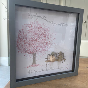 Framed Pebble Art - Grey block square frame 27.5cmWith a soft background image of a Tree full of pink flowers and a bench with two pebble friends sitting on it with the quote 'A good friend knows all your best stories... A best friend has lived them with you' 