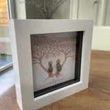 Mini Framed Pebble Art -White block square frame 12.5cmWith a soft background image of two trees full of leaves with two pebble people stood between the trees holding hands with the quote 'Love is all you need'