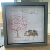 Framed Pebble Art - Grey block square frame 27.5cmWith a soft background image of a Tree full of pink flowers and a bench with two pebble friends sitting on it with the quote 'A good friend knows all your best stories... A best friend has lived them with you'