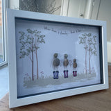 Framed Pebble Art - White block frame 31.5cmThe background image is tall trees on both sides with wild flowers growing and 3 Pebble people wearing colourful wellies holding hands with the flowers with the quote 'Where there is family...there is love' 