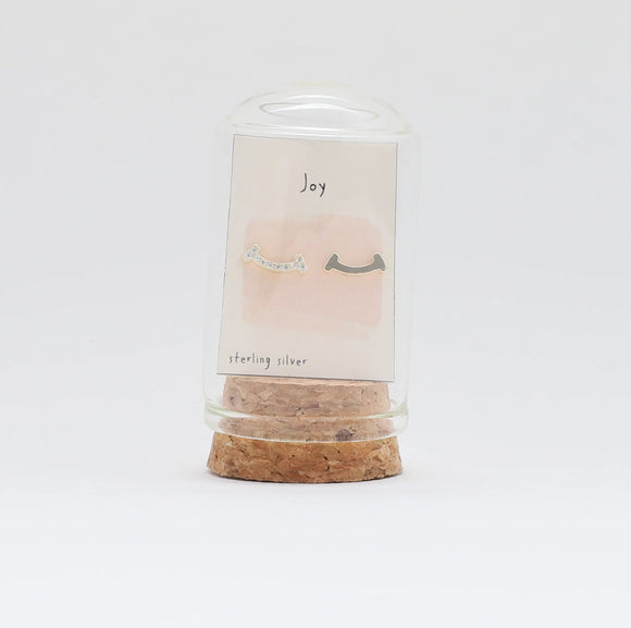 Asymmetrical smile shaped stud earrings presented in a bottle with a message that reads 